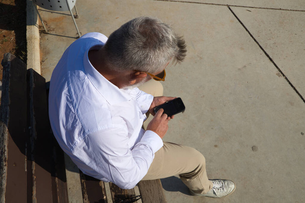 Handsome mature man with beard and grey hair is sitting thoughtfully on a park bench while consulting his mobile phone. The man is dressed in beige trousers, white shirt. Concept of old age. - 写真・画像