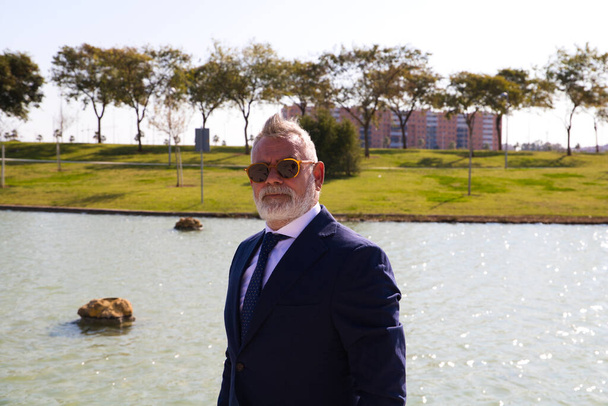 Handsome mature man, executive, grey hair, beard, sunglasses, jacket and tie, is in a park with an artificial lake posing for photo. Concept of mature man, grandfather, executive. - 写真・画像