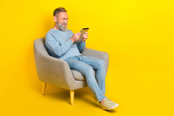 Full length portrait of aged person sit chair use tablet chatting typing isolated on yellow color background. - Fotó, kép