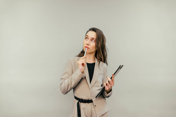 Pensive business woman in a suit stands on a beige background with a paper tablet and a pen in his hands, looks away with a serious face and thinks. - Photo, Image