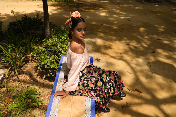 Piękna nastoletnia tancerka flamenco ubrana w kwiecistą spódnicę z falbankami i kwiatami we włosach siedzi na ławce w parku, uśmiechnięta i pozująca do zdjęć. - Zdjęcie, obraz