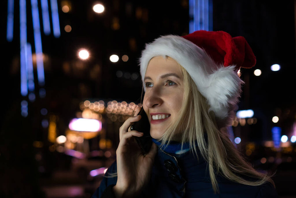 Молодая улыбающаяся блондинка в шляпе Санта Клауса разговаривает по телефону на освещенной улице ночного города. - Фото, изображение