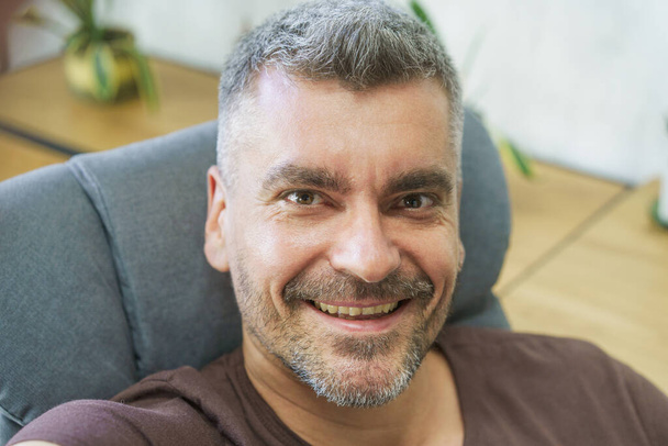 Handsome man in casual sitting in office chair, smiling making selfie using his smartphone in home or office interior. Scruffy face man making self portrait or video call in home interior.  - Fotografie, Obrázek