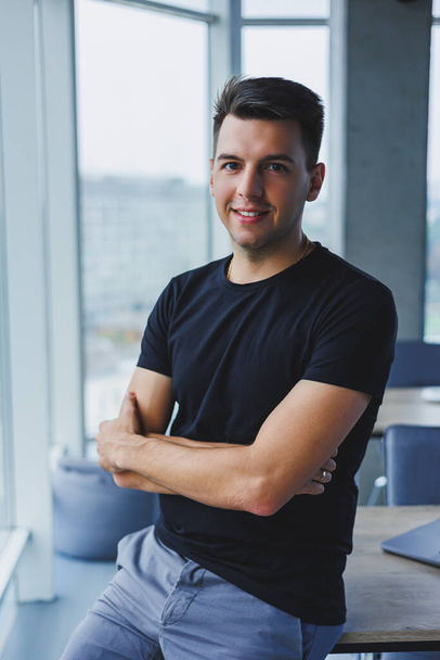 Portrait of a smiling young man in a black t-shirt against the background of a modern office. Stylish guy in casual clothes - Фото, изображение