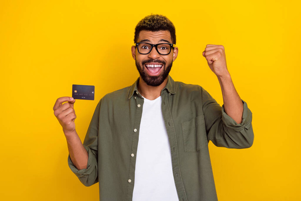 Photo of pretty lucky man wear khaki shirt spectacles rising fist holding credit card isolated yellow color background. - Foto, immagini
