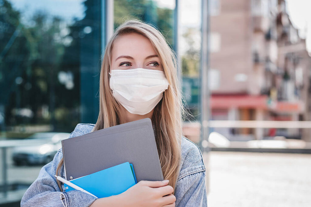 Quarantine coronavirus concept while studying. Caucasian blonde student girl wearing a protective medical mask holds notebook folders and look away. Close up portrait - Photo, Image