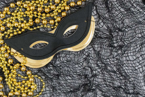 maskerade of Halloween achtergrond met een zwarte kat stijl masker en een metalen gouden masker en gouden kralen op een netting achtergrond - Foto, afbeelding