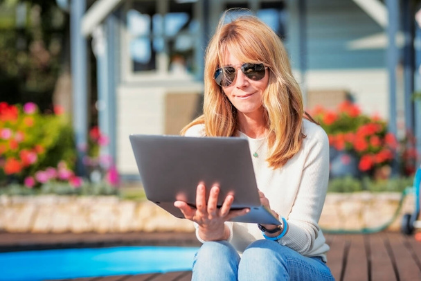 Attractive middle aged woman using laptop and having video call while sitting at the backyard at home. - Foto, immagini