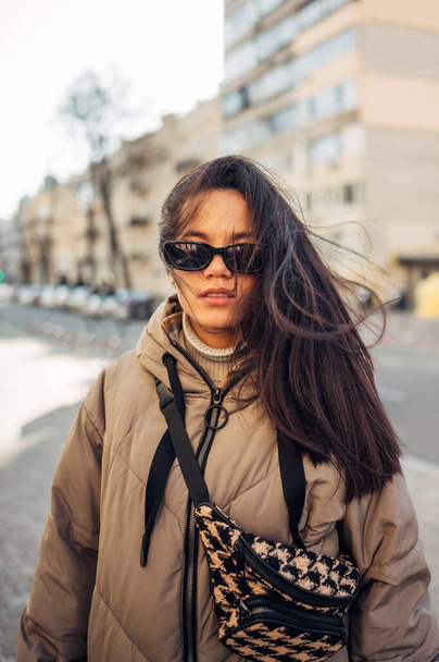 Attractive Hispanic woman in a jacket and sunglasses stands on the street and looks at the camera with a serious face. Vertical. - Fotó, kép