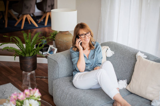 Happy middle aged woman wearing casual clothes and relaxing on a couch while having a call.  - Foto, Bild