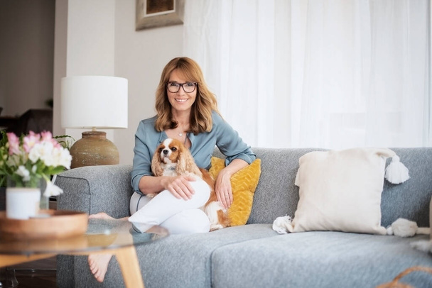 Shot of happy middle aged woman relaxing on the sofa at home with her cavalier puppy. - Foto, Imagen