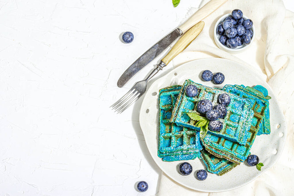 Hausgemachte belgische Waffeln mit Blaubeeren. Glutenfreies blaues Dessert, frisches Obst und Minze. Trendy hartes Licht, dunkler Schatten. Weißer Gipshintergrund, Draufsicht - Foto, Bild