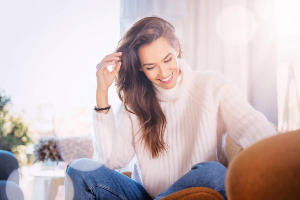 Shot of cheerful woman sitting in an armchair at home. Attractive woman laughing and wearing sweater while relaxing at home.  - Zdjęcie, obraz