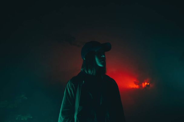 Woman in casual clothes and cap stands at night in a foggy park in green light on a background of red rays, looking to the side. - Фото, изображение