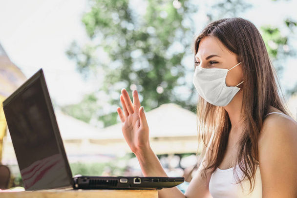 Eine junge Frau mit medizinischer Maske spricht per Videoanruf. Junge Kaukasierin mit medizinischer Atemschutzmaske winkt am Laptop-Bildschirm - Foto, Bild