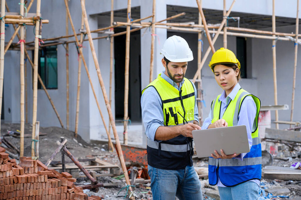 Young attractive construction man and woman in vest with helmet working with laptop, standing on building construction site. Home building project. Engineer foreman discusses with a coworker at workplace - Photo, image