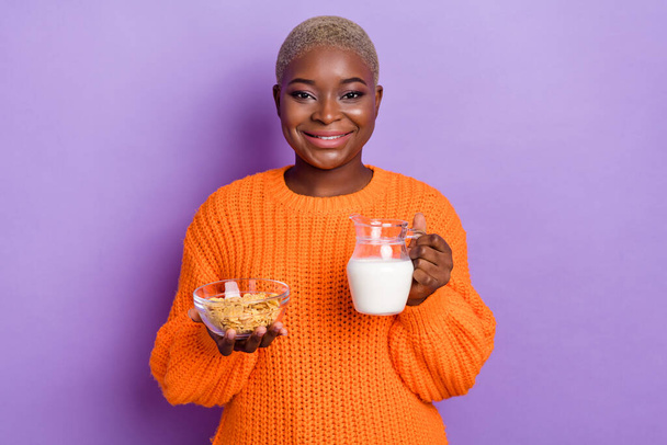 Φωτογραφία από ωραία νεαρή κυρία μαγειρεύουν δίαιτα μπολ δημητριακών γάλα κανάτα ντυμένο μοντέρνο φθινόπωρο πορτοκαλί πλεκτά πουλόβερ που απομονώνονται σε βιολετί χρώμα φόντο - Φωτογραφία, εικόνα