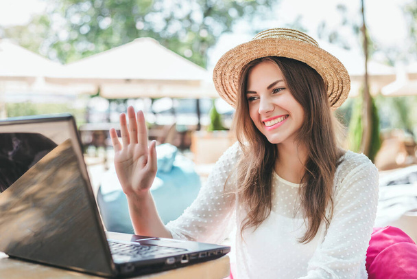 Jong freelance meisje glimlacht, zit aan een tafel op het strand, zwaait met haar hand op een laptop monitor en maakt videoconferentie aan haar vrienden, coronavirus, afstandswerk - Foto, afbeelding