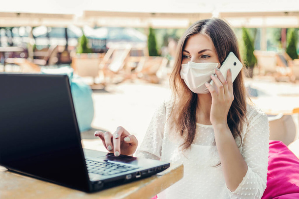 Trabajo a distancia. Una mujer con una máscara médica se sienta en la playa con un portátil en la mesa en un café y llama a un teléfono móvil, en busca de una vacante para un nuevo trabajo durante la cuarentena coronavirus - Foto, Imagen