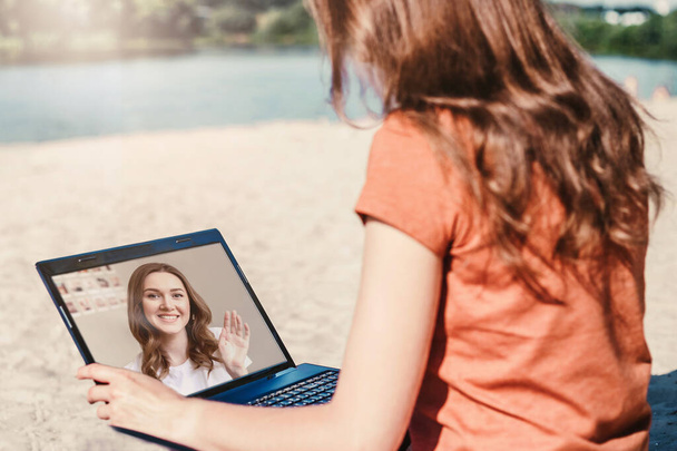 Due ragazze che parlano in videoconferenza su un computer portatile in webcam mentre sono sedute sulla spiaggia durante la quarantena coronavirus, covid-19, applicazione di videochiamate - Foto, immagini