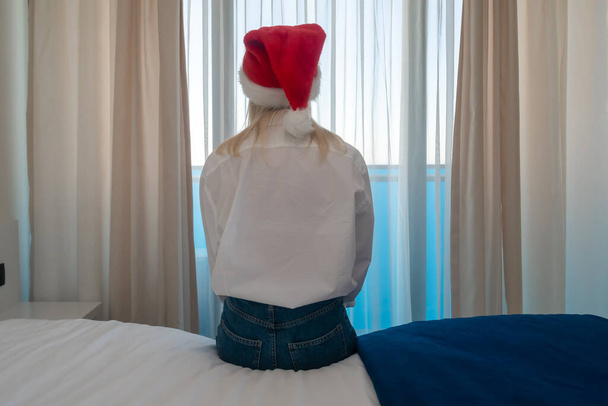 Sad young woman with drooping shoulders in a Santa Claus hat is sitting on the bed with her back to the camera and looking out the window - Zdjęcie, obraz