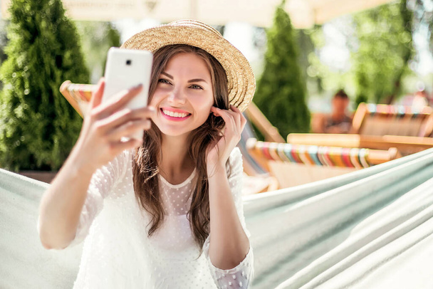 Mulher bonita jovem na moda sorrindo, tirando selfie no telefone celular, fazendo videoconferências durante as férias - Foto, Imagem