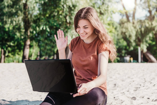 Brown-haired girl sitting on the beach with a laptop and waving her hand at the monitor screen, video conference, calls via the Internet concept - Foto, Imagem