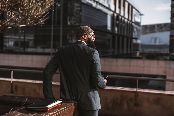 View from behind of a dapper bearded half-turned black man entrepreneur looking aside with a smartphone in his hand leaning against a street bed with an autumn tree; a copy space place on the right - Fotó, kép