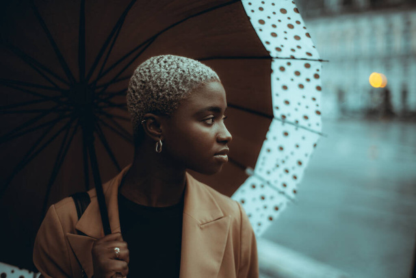A low-key portrait of a pensive cute youthful black female with short hair painted in white, looking aside while standing on a rainy street with a spotted umbrella; a copy space place on the right - Foto, Bild