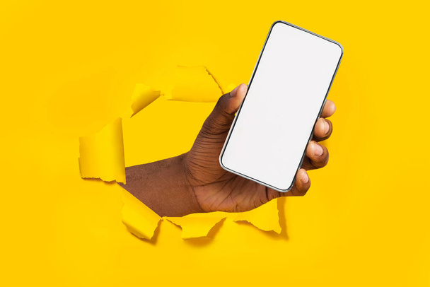 Αφροαμερικανοί άνδρες χέρι κρατώντας smartphone με λευκή άδεια οθόνη δείχνει τη συσκευή σπάζοντας μέσα από κίτρινο φύλλο χαρτιού. Οθόνη Gadget με χώρο αντιγραφής, μακιγιάρισμα - Φωτογραφία, εικόνα