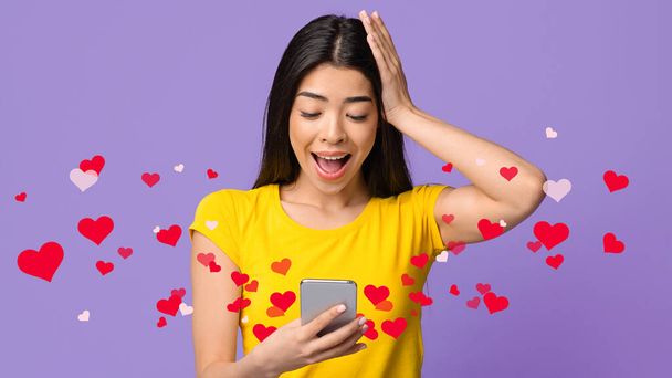 Úžasná asijská žena drží smartphone, dívá se na obrazovce zařízení s vzrušením, stojí s otevřenými ústy přes fialové studio pozadí s červenými srdci, emocionálně reaguje na milostné poselství, koláž - Fotografie, Obrázek