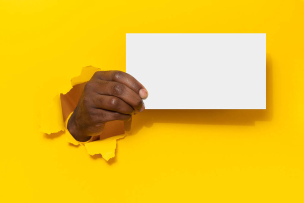 Schwarzer Mann hält leeres Plakat weißes Blatt durch Loch in gelbem Papierhintergrund, präsentiert leeren Raum für Ihren Entwurf oder Text, Attrappe - Foto, Bild