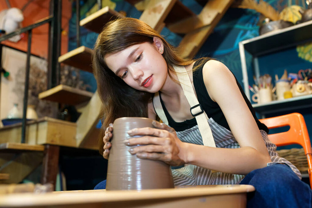 Close-up of concentrated beautiful craftswoman in apron sitting at pottery wheel and using craft tool while shaping wet clay vessel - Foto, Imagen