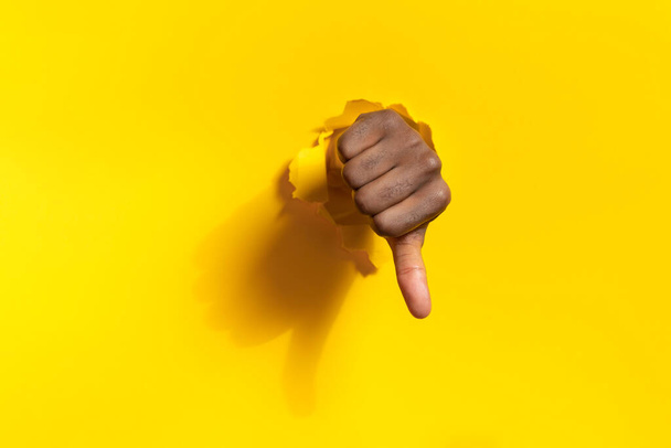 Mão masculina afro-americana mostrando polegar para baixo através de buraco rasgado em fundo de papel amarelo, espaço livre. Conceito de desagrado e gesto de desaprovação - Foto, Imagem