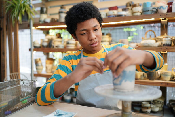 Молодой афро-подросток ручной гончар делает глиняную вазу в керамической мастерской - Фото, изображение