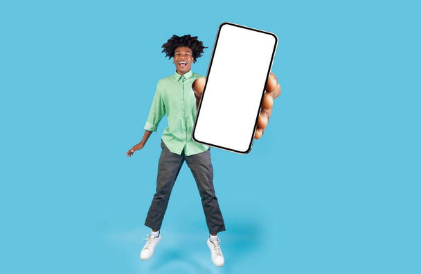 Fröhlicher schwarzer Teenie-Typ demonstriert großen leeren Smartphone-Bildschirm, während er über blauen Studiohintergrund springt, junge fröhliche afrikanisch-amerikanische Männer empfehlen Webseite oder mobile Anwendung, Attrappe - Foto, Bild