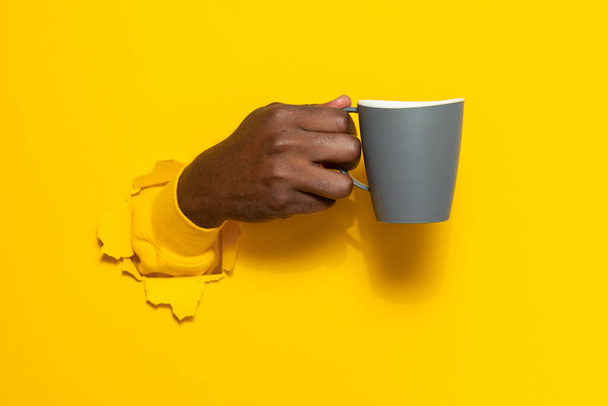 Schwarze männliche Hand, die einen Becher Kaffee durch zerrissenen gelben Papierhintergrund hält, Großaufnahme, Kopierraum. Eine Pause und ein erfrischendes Getränk - Foto, Bild