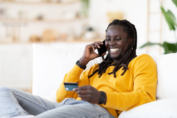 Online-pankkitoiminta. Iloinen Afrikkalainen Amerikkalainen Guy puhuu matkapuhelimella ja pitää luottokorttia, kun istuu mukavassa tuolissa kotona, hymyilevä musta mies tekee Internet-maksuja, vapaa tila - Valokuva, kuva