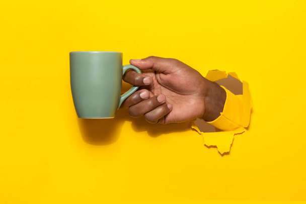 Αφροαμερικάνος που κρατάει ένα φλιτζάνι ζεστό ρόφημα μέσα από σκισμένο κίτρινο φόντο χαρτιού, κοντινό πλάνο, χώρος αντιγραφής. Άνδρας που κάνει διάλειμμα και πίνει καφέ - Φωτογραφία, εικόνα
