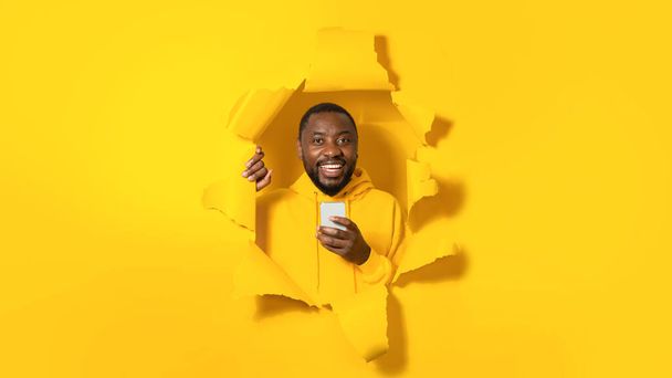 Happy black man holding modern cellular and smiling at camera, enjoying texting in social media, using mobile network services, posing in ripped torn yellow paper background - Photo, Image