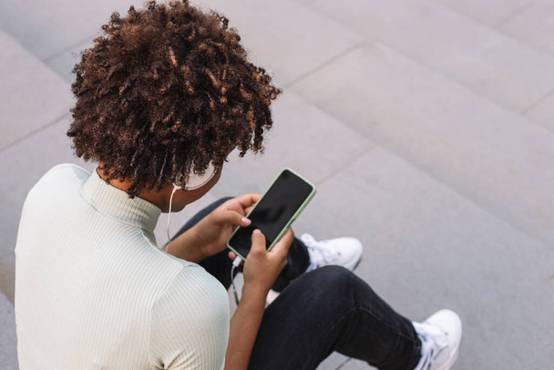 Νεαρή σγουρή γυναίκα δακτυλογραφεί ένα μήνυμα κειμένου μέσω του smartphone της, Λατίνα έφηβος με μπούκλες χρησιμοποιώντας το κινητό της τηλέφωνο - Φωτογραφία, εικόνα