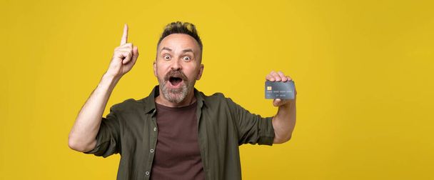 Astonished mature man notes with pointing up hand hoding bank card isolated on yellow background, studio portrait. People sincere excited emotions lifestyle and finance concept. - Foto, afbeelding