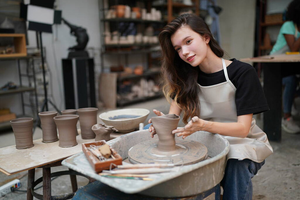 Gros plan d'une belle artisan concentrée dans un tablier assis à la roue de poterie et utilisant un outil artisanal tout en façonnant un récipient en argile humide - Photo, image