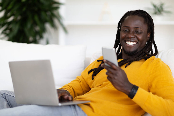 Muotokuva hymyilevä musta kaveri käyttää kannettavaa tietokonetta ja lukeminen viesti älypuhelimella lepää sohvalla kotona, onnellinen Afrikkalainen amerikkalainen mies nauttii nykyaikaisesta teknologiasta ja online-viestinnästä, lähikuva - Valokuva, kuva