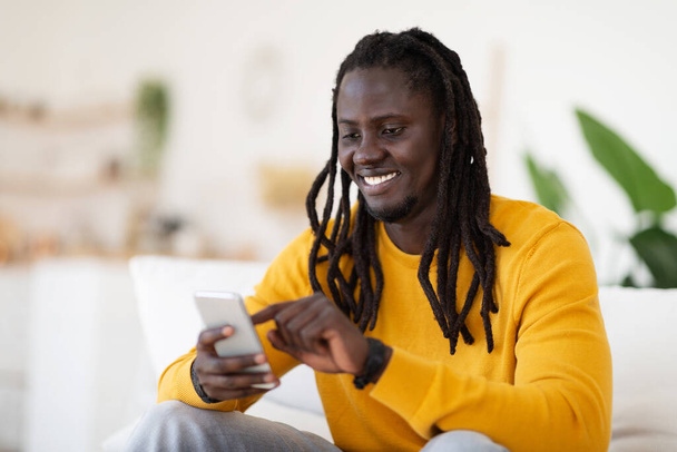 Цифровой досуг. Улыбающийся черный молодой человек просматривает социальные сети на смартфоне, расслабляясь дома, счастливый афроамериканец с мобильным телефоном в руках, сидя на диване в гостиной - Фото, изображение