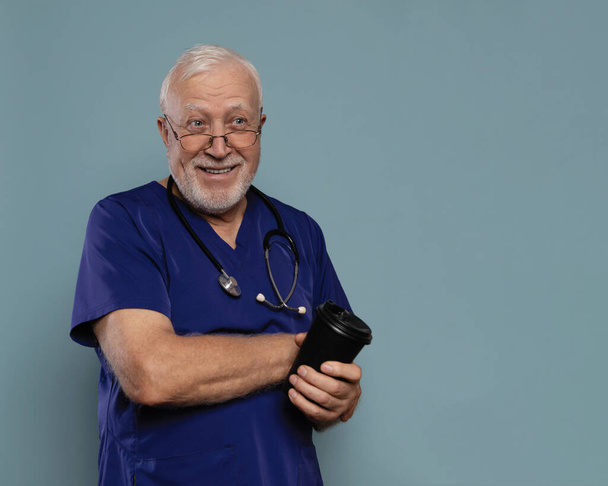 Charismatischer älterer Herr, Arzt, stehend, Brille mit Stethoskop und Kaffee und lächelnd, in medizinischer Kleidung auf blauem Hintergrund, familienmedizinisches Konzept, Platz für Text - Foto, Bild