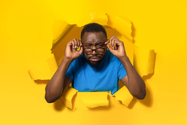 Black man with poor eyesight wearing eyeglasses and squinting eyes, looking at camera through hole in torn yellow paper. Healthcare, vision problems - Photo, Image