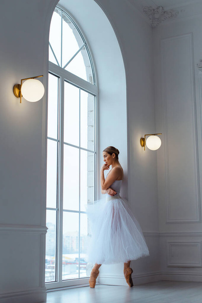 Балерина в длинной белой юбке тюль в комнате на фоне большого окна, поза на цыпочках - Фото, изображение