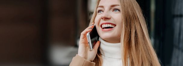 Young blonde Caucasian woman in a beige coat talking on a mobile phone, smiling and looking up against the background of an office building. Corporate employee. Happy female IT-specialist. Copy space - Photo, image