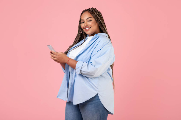Felice nero grassoccio donna utilizzando smartphone sms nei social media sorridendo alla fotocamera in posa su sfondo rosa in studio. Pubblicità nuova applicazione mobile - Foto, immagini